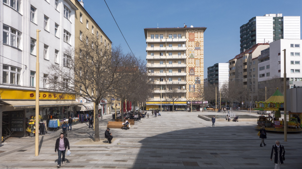 Ein belebter Stadtplatz mit Menschen, die gehen, auf Bänken sitzen und stehen, umgeben von Bäumen, Gebäuden und einem Karussell im Hintergrund unter einem sichtbaren Himmel, mit Strommasten und Drähten entlang der Straße.