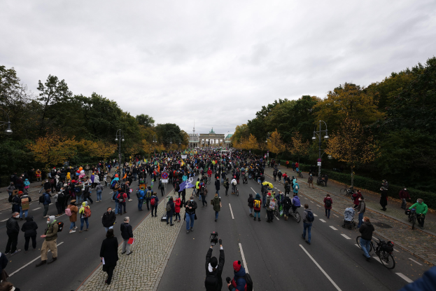Große Gruppe von Menschen geht eine Straße mit Bäumen und Laternen entlang, hält Kameras in der Hand, während sie an einer Protestdemo in Berlin teilnimmt, mit einem Gebäude und einem klaren Himmel im Hintergrund.