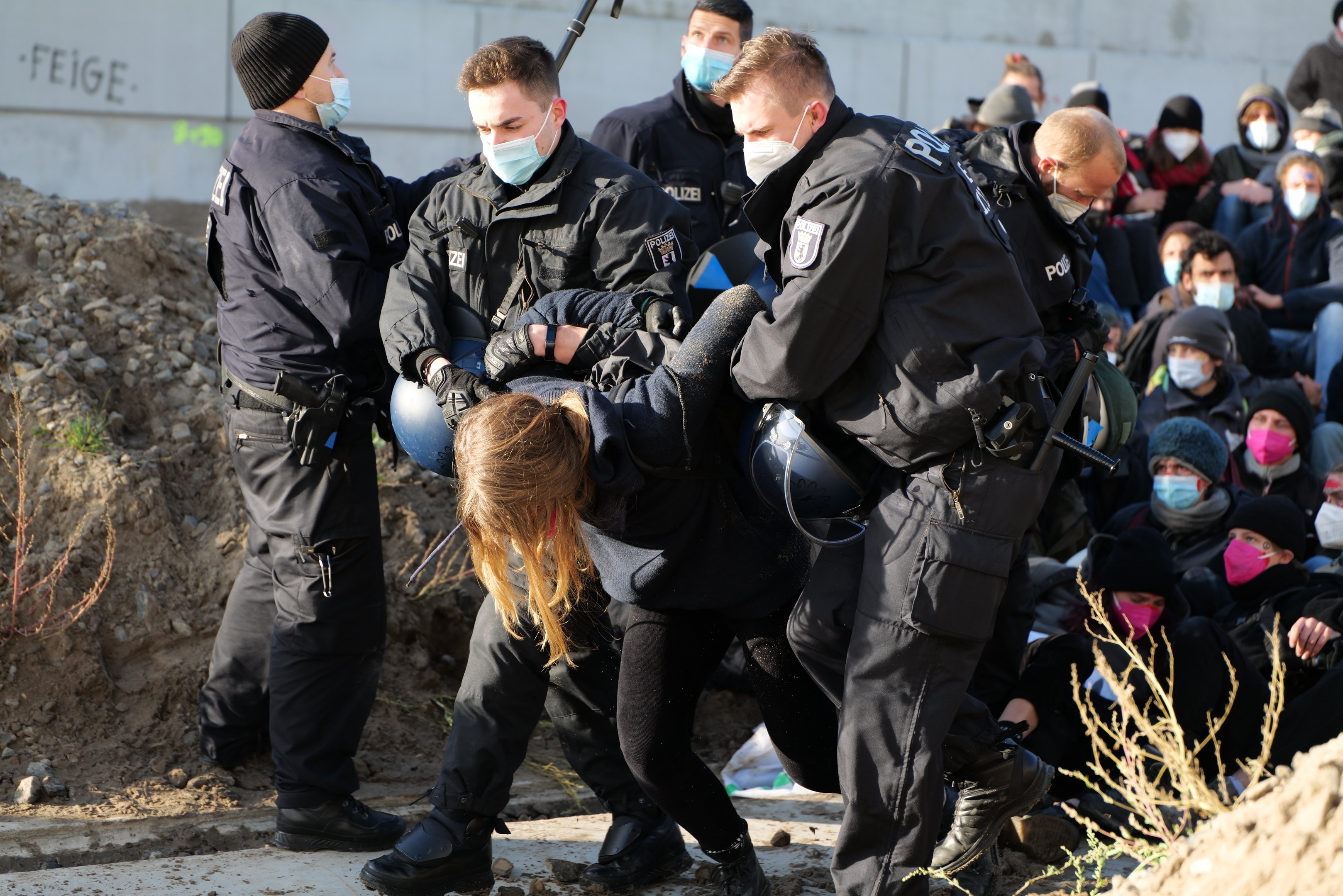Gruppe maskierter Personen in Kappen, einige bewaffnet, umringen eine Frau, die von der Polizei in einer städtischen Umgebung mit Pflanzen, Steinen und protestbezogenen Graffiti an einer Wand begleitet wird.