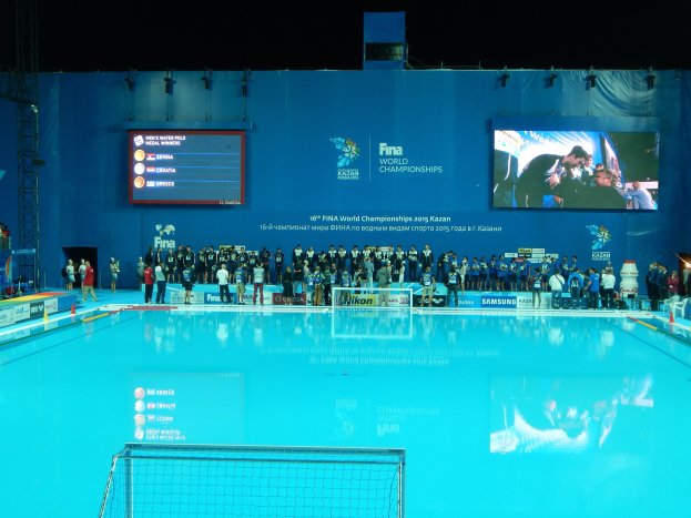 Gruppe von Menschen um einen Hallenbadpool mit Netz am Boden herumstehend mit einer Wand im Hintergrund, die "FINA World Championships 2015 Kazan"-Text und Logos, Trägern und Beleuchtung anzeigt.