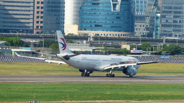 Ein großes Passagierflugzeug startet von einer Flughafenpiste mit umgebenem Gras, einem Zaun, Bäumen, Straßenlaternen, Schildern, einer Brücke, Gebäuden mit Fenstern und einem bewölkten Himmel.