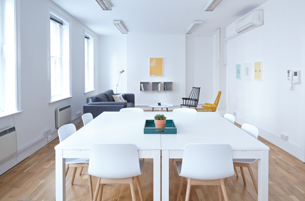 Innenansicht eines Apartments in Berlin mit einem weißen Tisch mit einer Topfpflanze, Stühlen, einem Sofa mit Kissen, einer Lampe, Regalen mit Gegenständen, einer Klimaanlage, Fotorahmen an der Wand, Fenstern und Deckenleuchten.