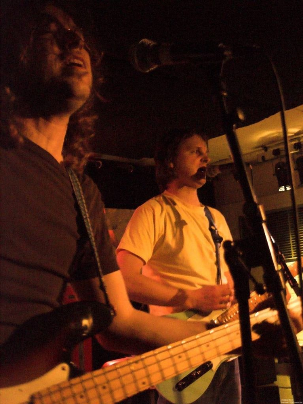 Ein Musikkonzert mit zwei Sängern: einer in einem weißen Hemd mit einer grünen Gitarre und einer in einem schwarzen Hemd mit Brille und einer schwarzen Gitarre, beide performen mit Mikrofonen; Bühnenlicht und zusätzliche Instrumente sind im Hintergrund sichtbar.