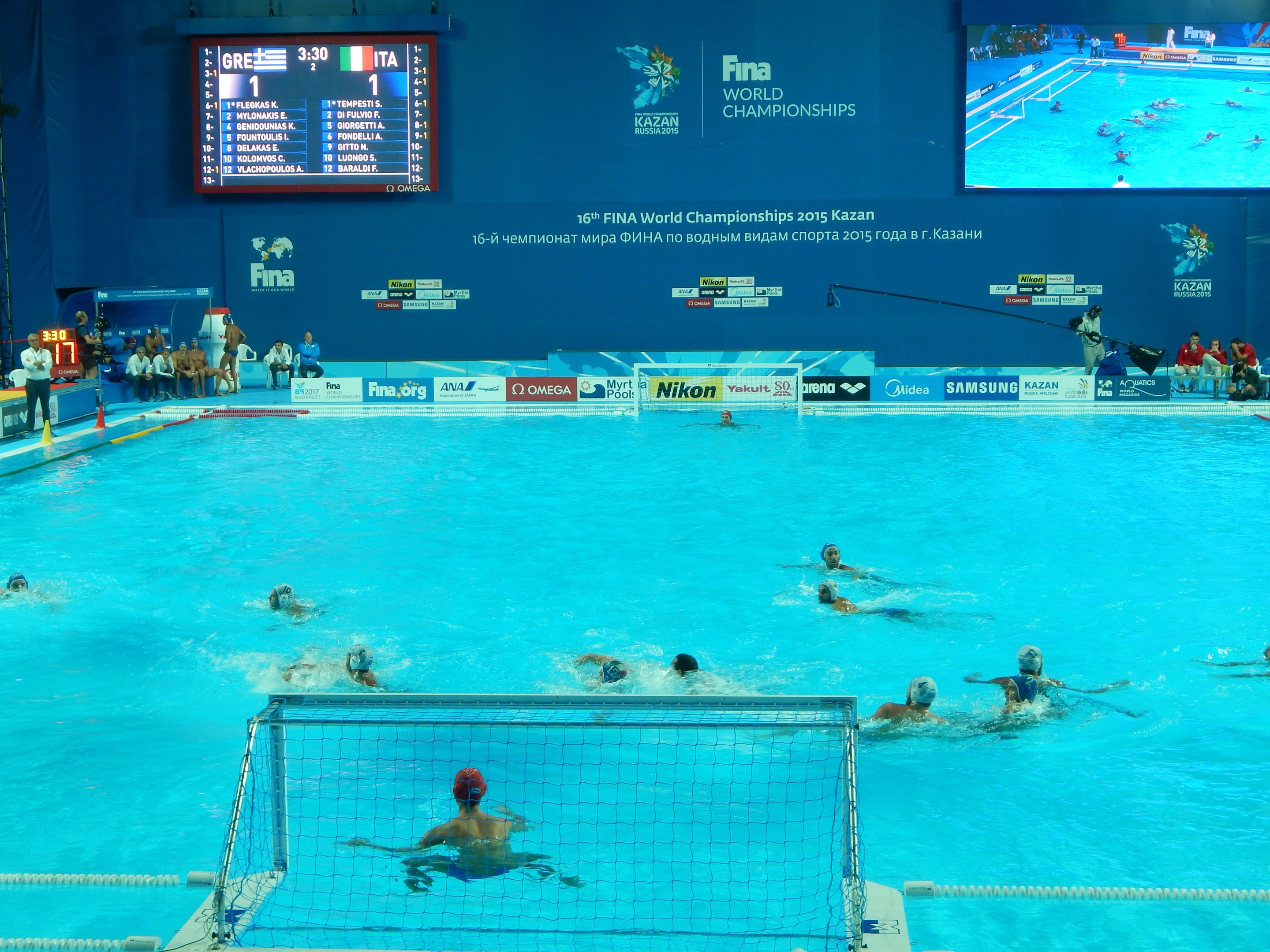 Wasserballspieler bei den FINA-Weltmeisterschaften 2016 in Kazan im Pool gegeneinander antretend, mit Zuschauern auf der linken Seite und einem Anzeigebrett mit Event-Logos im Hintergrund.