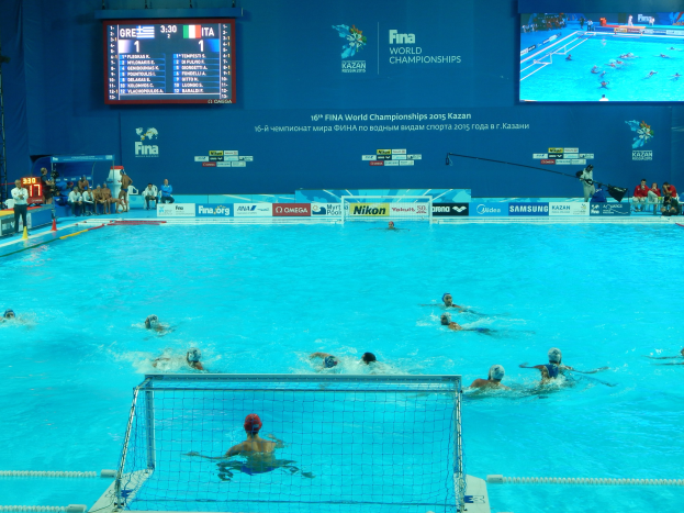 Wasserballspieler bei den FINA-Weltmeisterschaften 2016 in Kazan im Pool gegeneinander antretend, mit Zuschauern auf der linken Seite und einem Anzeigebrett mit Event-Logos im Hintergrund.