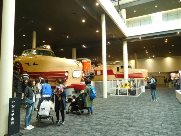 Eine Gruppe von Menschen, die eine Bahnausstellung in einem Museum mit Säulen, Geländern und Deckenleuchten betrachtet, einige tragen Taschen oder Kinderwagen, und ein Textschild links.