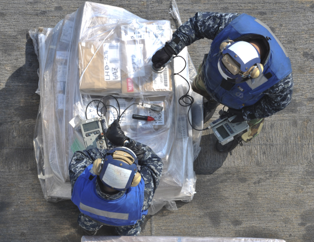 Zwei Männer in blauen Uniformen und Helmen arbeiten an einem großen Drohnenmodell und halten verschiedene Gegenstände in den Händen, mit einer Plastikabdeckung auf dem Boden.