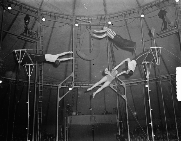 Ein Schwarz-Weiß-Foto von Akrobaten, die in einem Zirkuszelt performen, umgeben von Stangen, Leitern und Lichtern, mit einer Zuschauermenge unten.