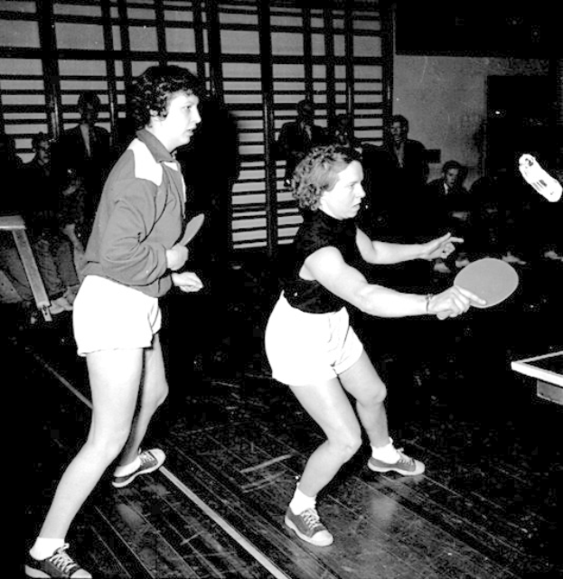 Zwei Frauen beim Tischtennis auf einem Court, jede hält einen Schläger in der Hand, mit Zuschauern im Hintergrund und einem Tischtennis-Tisch auf der rechten Seite, in Schwarz-Weiß.