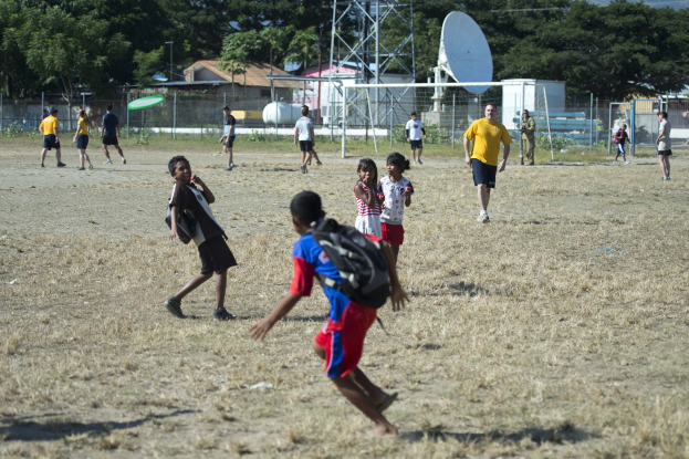 Kinder spielen Fußball auf einem eingezäunten Feld mit Häusern, Hügeln und einem bewölkten Himmel im Hintergrund; einige Kinder tragen Taschen.