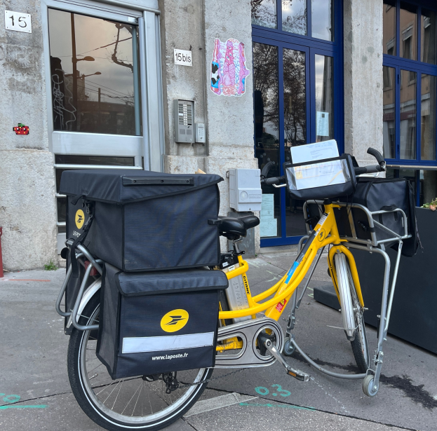 Ein gelbes Lieferrad mit mehreren Taschen am Hinterrad, das vor einem Gebäude mit Glastüren und Fenstern geparkt ist.