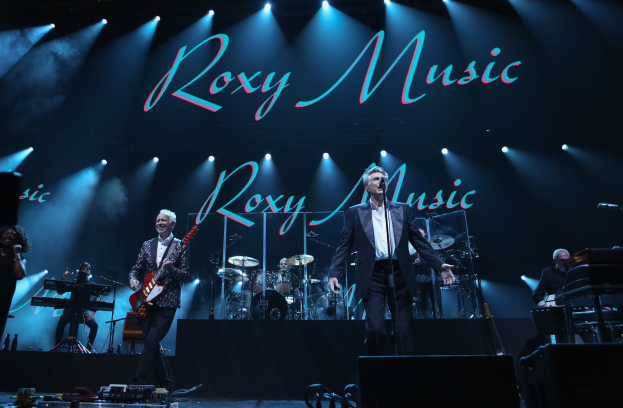 Eine Live-Band-Aufführung auf der Bühne mit Instrumenten, Mikrofonen und einem Hintergrund, der 'Roxy Music - Live at the O2 Arena, London' anzeigt.