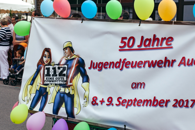 Gruppe von Menschen auf einer Straße mit Kinderwagen, mit einem Banner mit der Aufschrift "50 Jahre Jugendfeuerwehr August 2017", Ballons, einem Zelt, einem Zaun, Bäumen und einem Hintergrundgebäude.