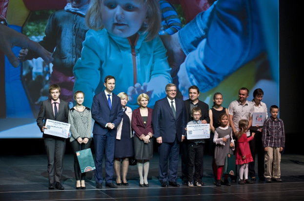Eine Gruppe von Menschen auf einer Bühne, einige halten Taschen und Urkunden, im Hintergrund ein Bildschirm mit einem Bild eines Kindes und anderer Menschen.