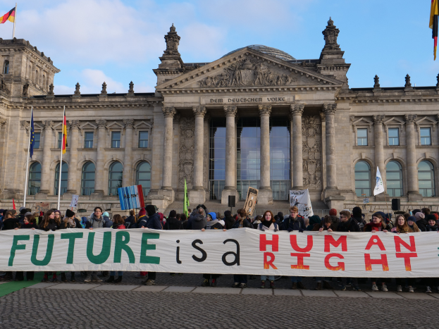 Eine Gruppe von Menschen hält ein Transparent mit der Aufschrift "Zukunft ist ein Menschenrecht" vor dem Reichstagsgebäude in Berlin, Deutschland, das mit Fenstern, Säulen und Bögen verziert ist und von Fahnenstangen umgeben ist, unter einem bewölkten Himmel.