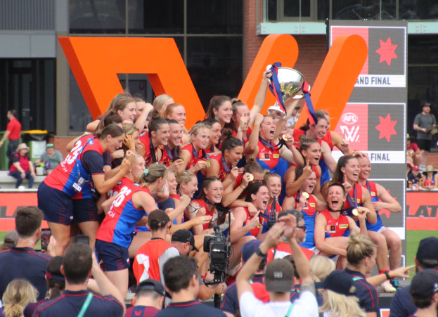 Eine Gruppe von Frauen-AFL-Spielerinnen in blauen und roten Uniformen feiert mit einem Pokal vor einem jubelnden Publikum, mit Menschen, Texttafeln und einem Gebäude mit Glasfenstern im Hintergrund auf einem grünen Feld.