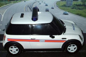 Weißer Polizeiwagen mit "Polizei"-Aufschrift auf einer Straße außerhalb einer Stadt, umgeben von fahrendem Verkehr, Gras und Bäumen auf beiden Seiten.