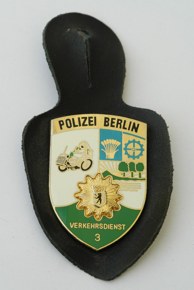 Ein schildförmiges Polizeibadged mit "Polizei Berlin" in schwarzer Schrift, das einen blauen Hintergrund, einen weißen Stern und einen weißen Rand hat und auf einer Oberfläche liegt.