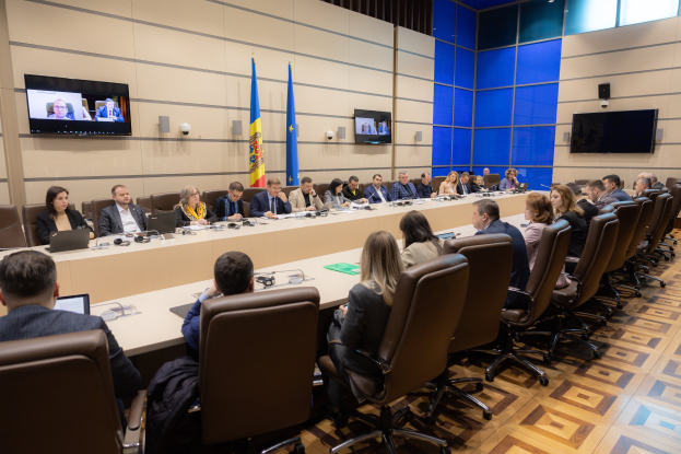 Große Gruppe von Menschen sitzt um einen langen Tisch mit Laptops, Mikrofonen, Papieren und anderen Gegenständen während einer Sitzung des Europäischen Rates für Menschenrechte, mit drei Flaggen, zwei Fernsehern und Deckenleuchten im Hintergrund.