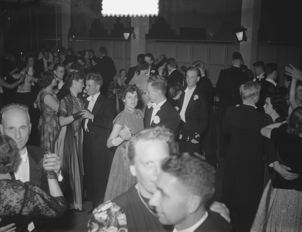 Gruppe von Menschen, die auf einer Party in einer Schwarz-Wei├č-Einstellung tanzen, mit M├Ąnnern und Frauen verschiedenen Alters, einige halten Gl├Ąser, und eine beleuchtete Wand im Hintergrund.