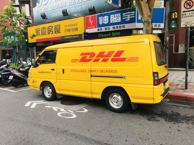 Ein gelber DHL-Lieferwagen parkt am Straßenrand, umgeben von Gebäuden, Bäumen, Schildern, Pfählen, Verkehrszeichen und einer Person auf dem Gehweg.