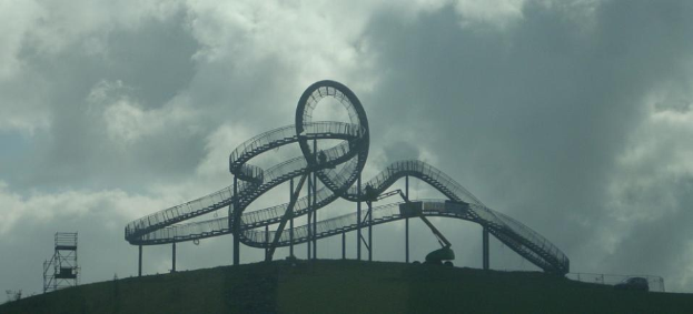 Eine Achterbahn auf dem Höhepunkt eines Hügels bei einem bewölkten Himmel, umgeben von Stützpfählen und -strukturen.