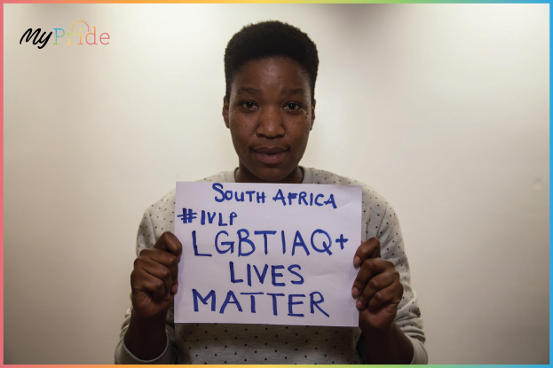 Eine Frau in einem weißen Hemd hält ein Schild mit der Aufschrift "LGBTQ+ Lives Matter" vor einer Wand hoch, mit einem entschlossenen Gesichtsausdruck.