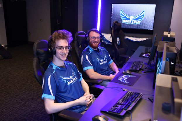 Zwei Männer mit Headsets sitzen an einem Schreibtisch mit Monitoren, Tastaturen und anderem Equipment und lächeln, während sie ein Spiel in einem Raum mit einem Bildschirm und Lichtern im Hintergrund spielen.