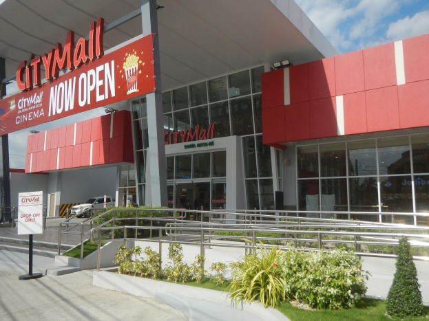 Außenansicht eines neu eröffneten Stadtzentrums mit einem "Stadtzentrum Eröffnet"-Schild, umgeben von Grün, einem Metallzaun, Menschen, einem Auto, Pfählen, einer Fahne, Lichtern und einem bewölkten Himmel.