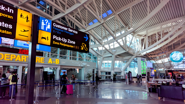 Ein Blick ins Innere eines Flughafen-Terminals mit Menschen, die gehen, einige mit Gepäck, und Pfosten mit Bändern und Texttafeln; Sofas und Tische mit Gegenständen auf der rechten Seite und ein Gebäude mit Glaswänden und Deckenleuchten im Hintergrund.