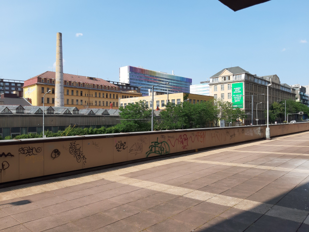 Stadtstraßenansicht mit Graffiti-bedeckter Wand, Bäumen, Straßenlaternen, Hinweisschild, Fahrzeugen, Gebäuden mit Fenstern, Schornstein und bewölktem Himmel.