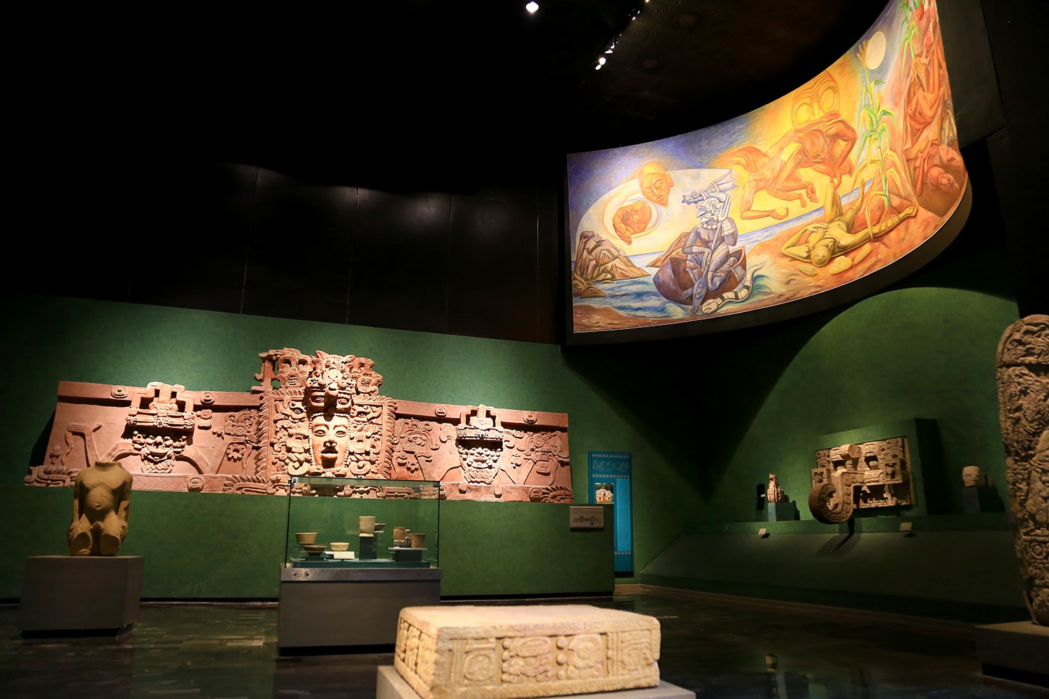 Ein Museumsraum mit verschiedenen Artefakten, darunter Skulpturen, Tischobjekte, eine Texttafel, eine Wandmalerei und Deckenbeleuchtung im Anthropologie-Museum von Mexiko-Stadt.