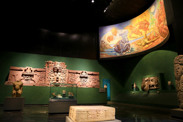 Ein Museumsraum mit verschiedenen Artefakten, darunter Skulpturen, Tischobjekte, eine Texttafel, eine Wandmalerei und Deckenbeleuchtung im Anthropologie-Museum von Mexiko-Stadt.
