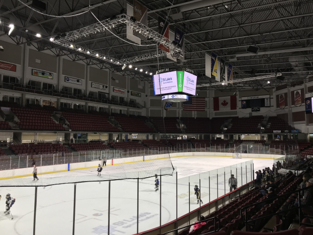 Hockey-Spiel in einer großen Arena mit einem zentralen Netz, Zuschauern auf beiden Seiten, Deckenbeleuchtung und Trägern sowie Bannern an den Wänden, die "St. Louis Blues Arena" beschriftet sind.