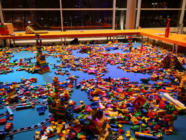 Ein großer Raum mit farbigen Bauklötzen, die auf dem Boden verstreut sind, umgeben von Tischen und Säulen, durch Glasfenster mit Menschen und Ausstellungsstücken in einem Lego-Museum.
