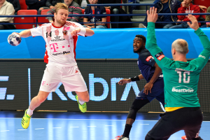 Eine Gruppe von Männern, die Handball auf einem Platz spielen, mit einem Ball in der Mitte der Aktion, whärend Zuschauer von Stühlen und Gelöndern im Hintergrund zuschauen und eine Tafel mit 'Futsal-Weltmeisterschaft 2019 - Bayern München vs. Paris Saint-Germain' anzeigt.