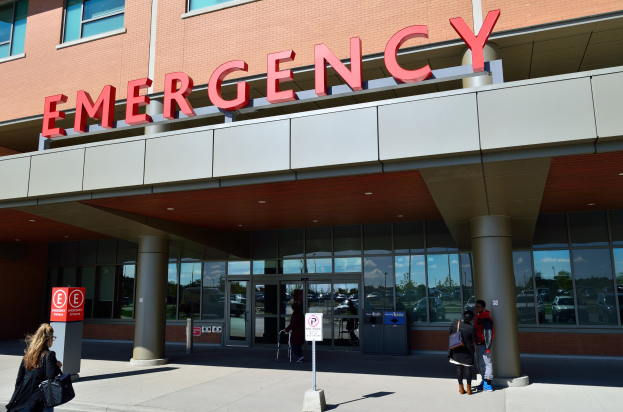 Eine Gruppe von Menschen vor einem Notfallgebäude mit Glaswänden und Säulen, einem Namensschild, Hinweistafeln mit Stangen und einem Mülleimer.