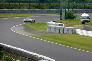 Ein Rennwagen fährt eine Rennstrecke hinunter, im Hintergrund ein Polizeiauto, umgeben von Grünflächen und Absperrungen, mit Text am unteren Bildrand.
