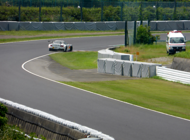 Ein Rennwagen fährt eine Rennstrecke hinunter, im Hintergrund ein Polizeiauto, umgeben von Grünflächen und Absperrungen, mit Text am unteren Bildrand.