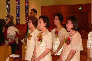 In der Mitte sind einige Frauen in Kleidern mit Blumen zu sehen, vor Bänken auf der linken Seite stehen einige Personen, ein Stück Papier auf einer Bank unten und eine Wand mit einer Tür oben.