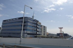 Ein großes Gebäude mit vielen Fenstern an der Spitze, das als Hauptquartier des Tokyo International Airport dient, umgeben von Straßenlaternen, Straßenlaternen, Kraftfahrzeugen, Absperrpollern, Schildern, Bäumen und einem bewölkten Himmel.