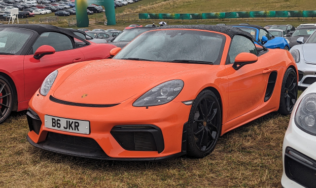 Roter Porsche 718 Boxster GTS auf dem Goodwood Festival of Speed mit Autos auf Gras, einem Banner, Bäumen und einem klaren blauen Himmel.