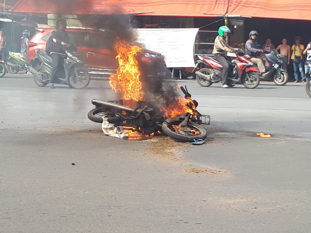 Motorrad in Flammen an der Straße mit einem Auto und einem Van im Hintergrund, umgeben von Menschen, einige tragen Helme, mit einer Fahne und einem Zelt in der Szene.
