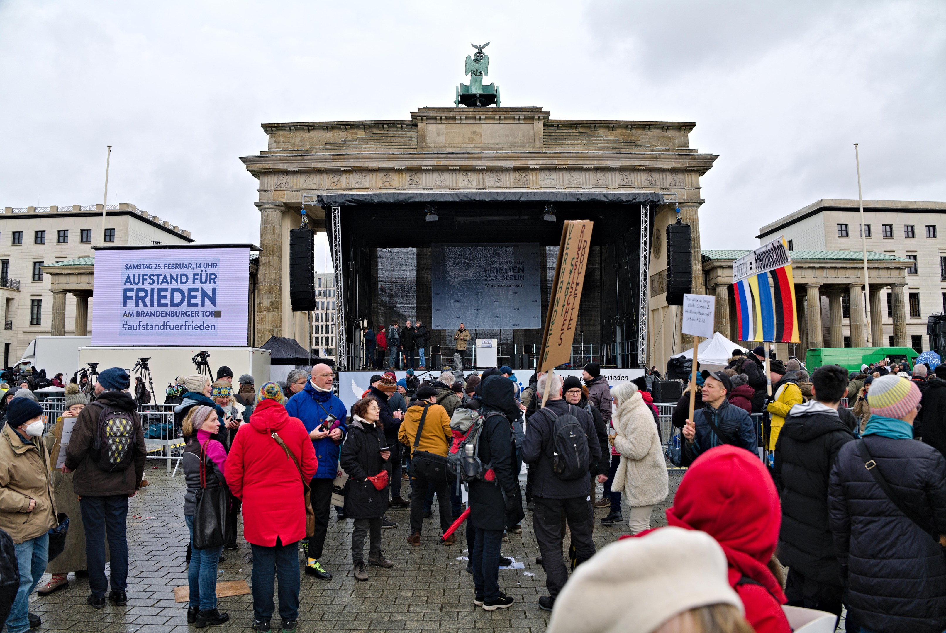 Eine Menschenmenge steht vor einem Gebäude mit einer Bühne, auf der Redner und ein Bildschirm zu sehen sind, umgeben von Fahnen und Transparenten mit Texten in Berlin während einer Demonstration.