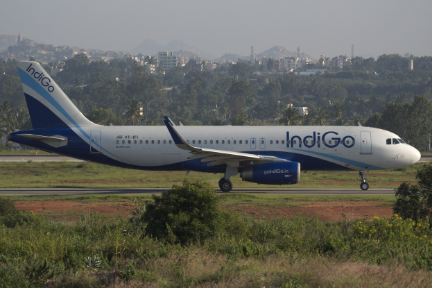 Indigo Airlines Airbus A320-200 auf dem Flughafen Mumbai mit Gras, Pflanzen, Geb├Ąuden, Türmen, Bergen und Himmel im Hintergrund.