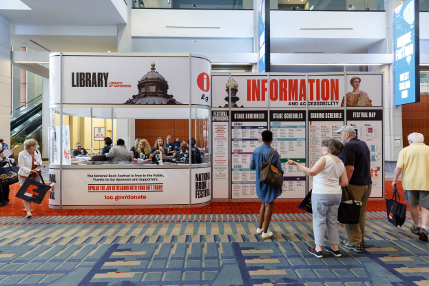 Eine Gruppe von Menschen auf dem Nationalen Buchfestival steht und sitzt in der Nähe einer Theke mit Bannern, einem Anzeigebildschirm, einer Treppe und Deckenleuchten.