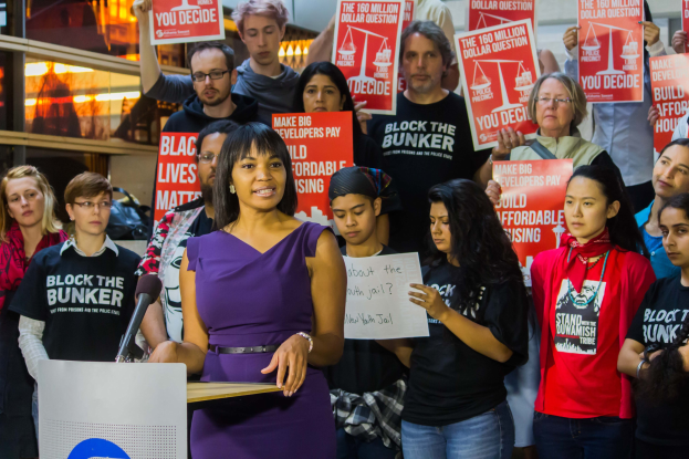 Eine Black Lives Matter-Aktivistin spricht an einem Podium mit Mikrofon vor einer Menge mit Schildern, mit Lichtern und Gegenständen im Hintergrund.
