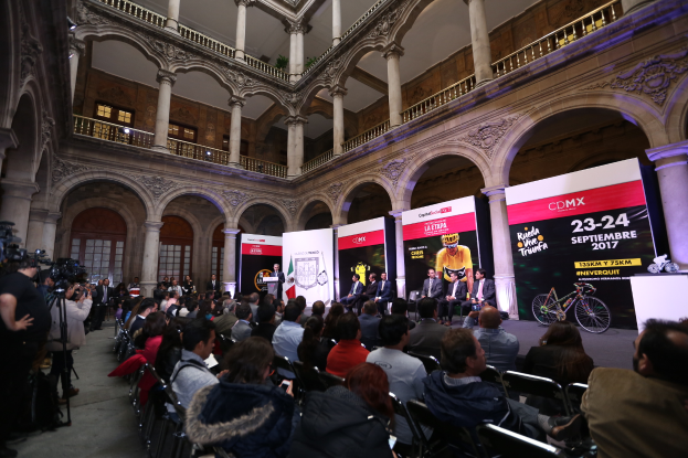 Eine Gruppe von Menschen sitzt an einem Tisch mit einem Podium und einem Mikrofon vor einer Menge, Kameras und Bannern, was auf eine Pressekonferenz für das CDMX 2017-Event hindeutet.
