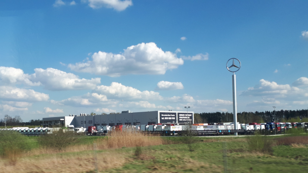 Eine Mercedes-Benz-Fabrik in Deutschland mit Fahrzeugen auf der Straße, umgeben von Gras, Pflanzen, Bäumen, Gebäuden und Polen, einschließlich einem mit einem Logo, unter einem bewölkten Himmel.