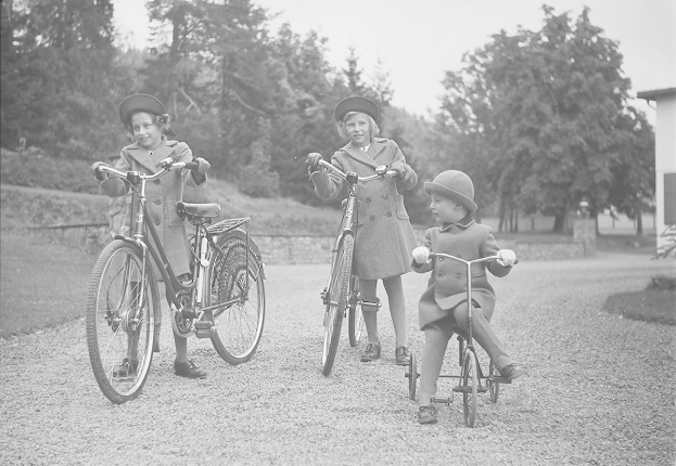 Drei Kinder auf Fahrrädern in einem Park, mit Mänteln und Hüten, mit Bäumen und einem Haus im Hintergrund, in Schwarz-Weiß.